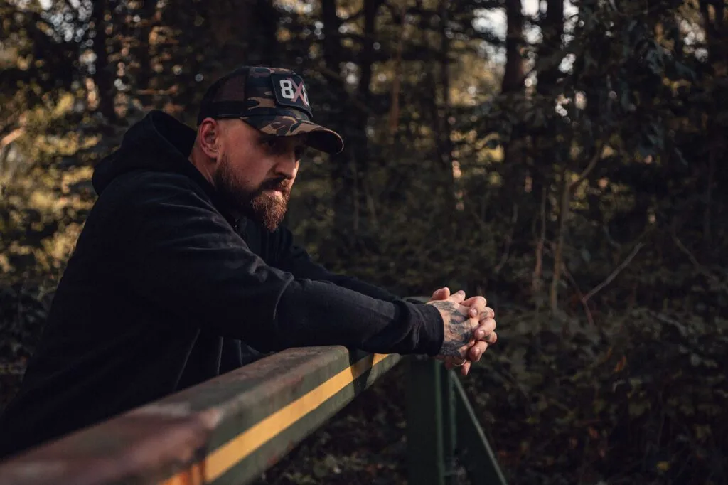 Man wearing a black 8x8 hoodie and camo trucker cap leaning on a woodland bridge.