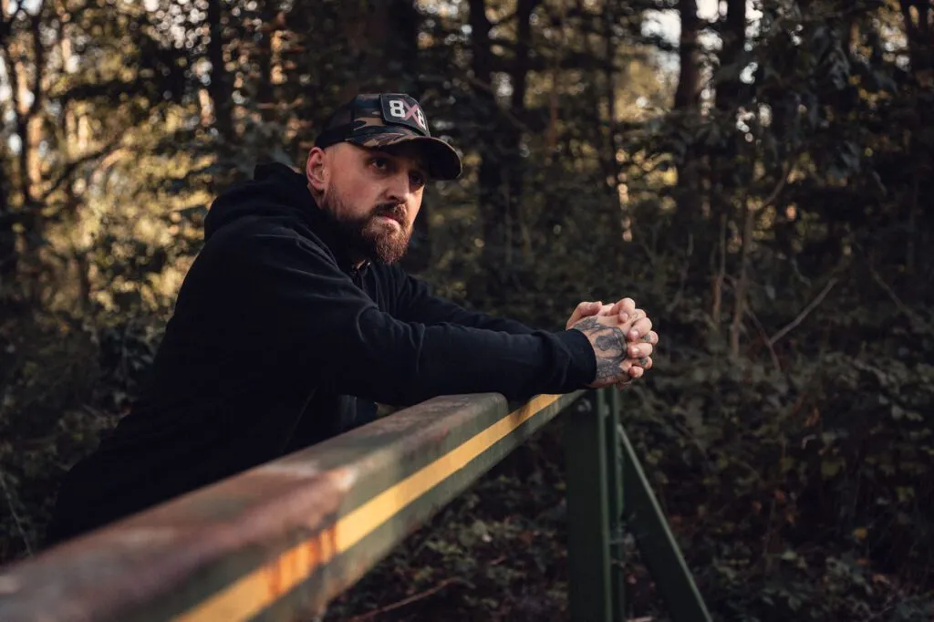 Man in a black 8x8 hoodie and camo trucker cap leaning on a woodland bridge railing.