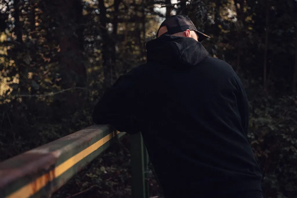 Rear view of a black 8x8 hoodie and camo trucker cap at a woodland bridge.