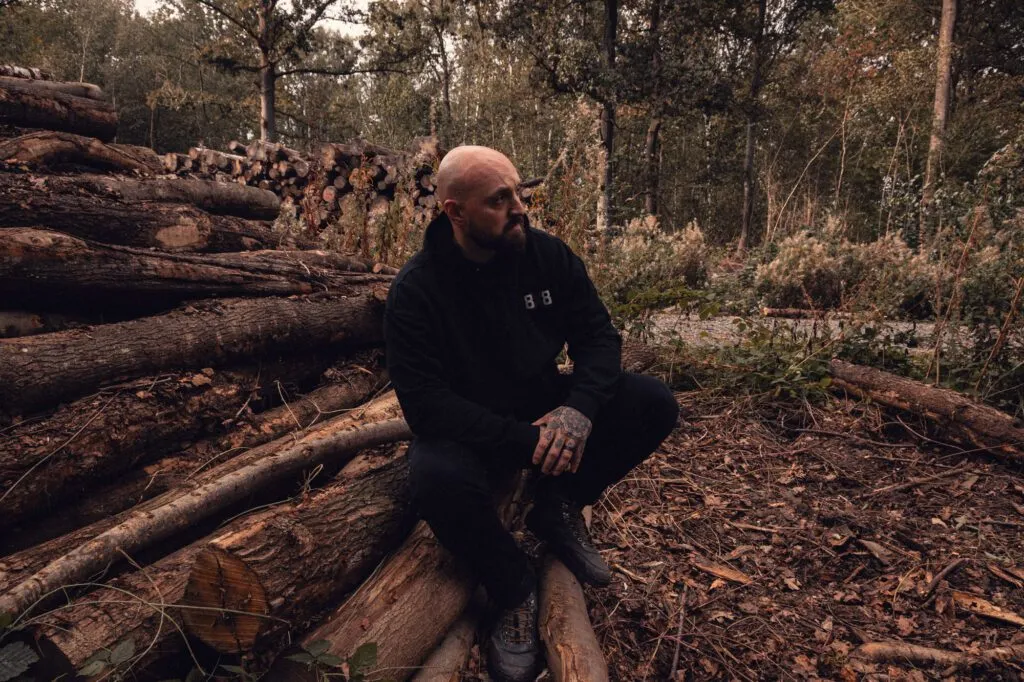 Black 8x8 hoodie worn while sitting beside a timber stack in woodland.