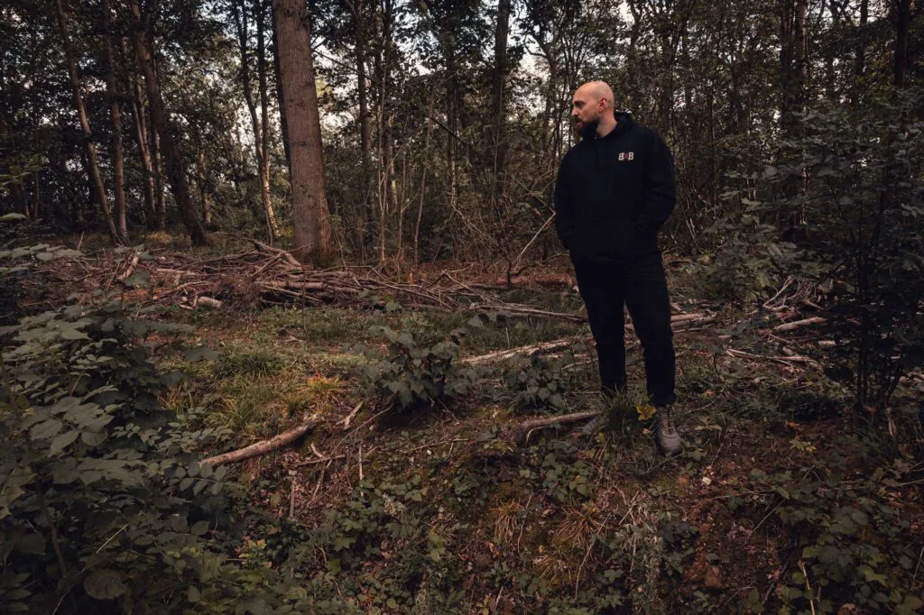 Man wearing a black 8x8 hoodie standing among trees in woodland.