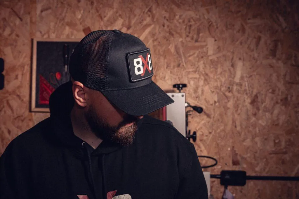 Lifestyle side view of the black 8x8 trucker cap worn in a workshop.