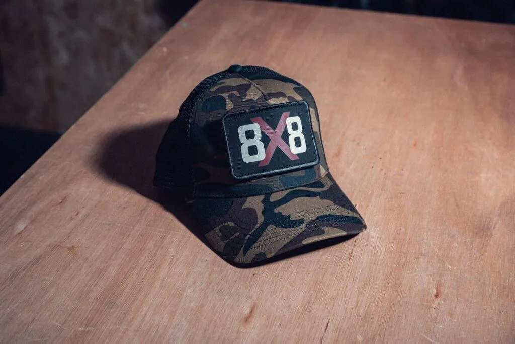8x8 camo trucker cap with logo patch on a wooden workshop bench.