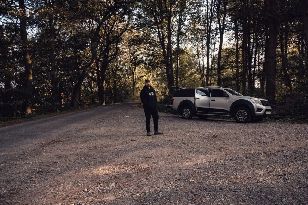 Man wearing the black 8x8 OG hoodie beside a pickup on a woodland track.