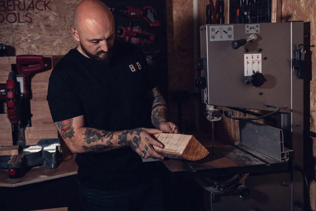 Man wearing the black 8x8 OG T-shirt holding timber beside a bandsaw.