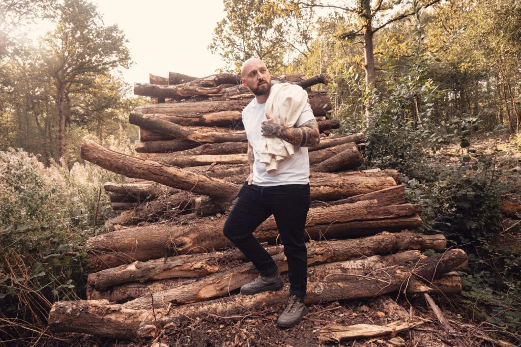 Man wearing a white 8x8 T-shirt beside a log pile with a hoodie over his shoulder.